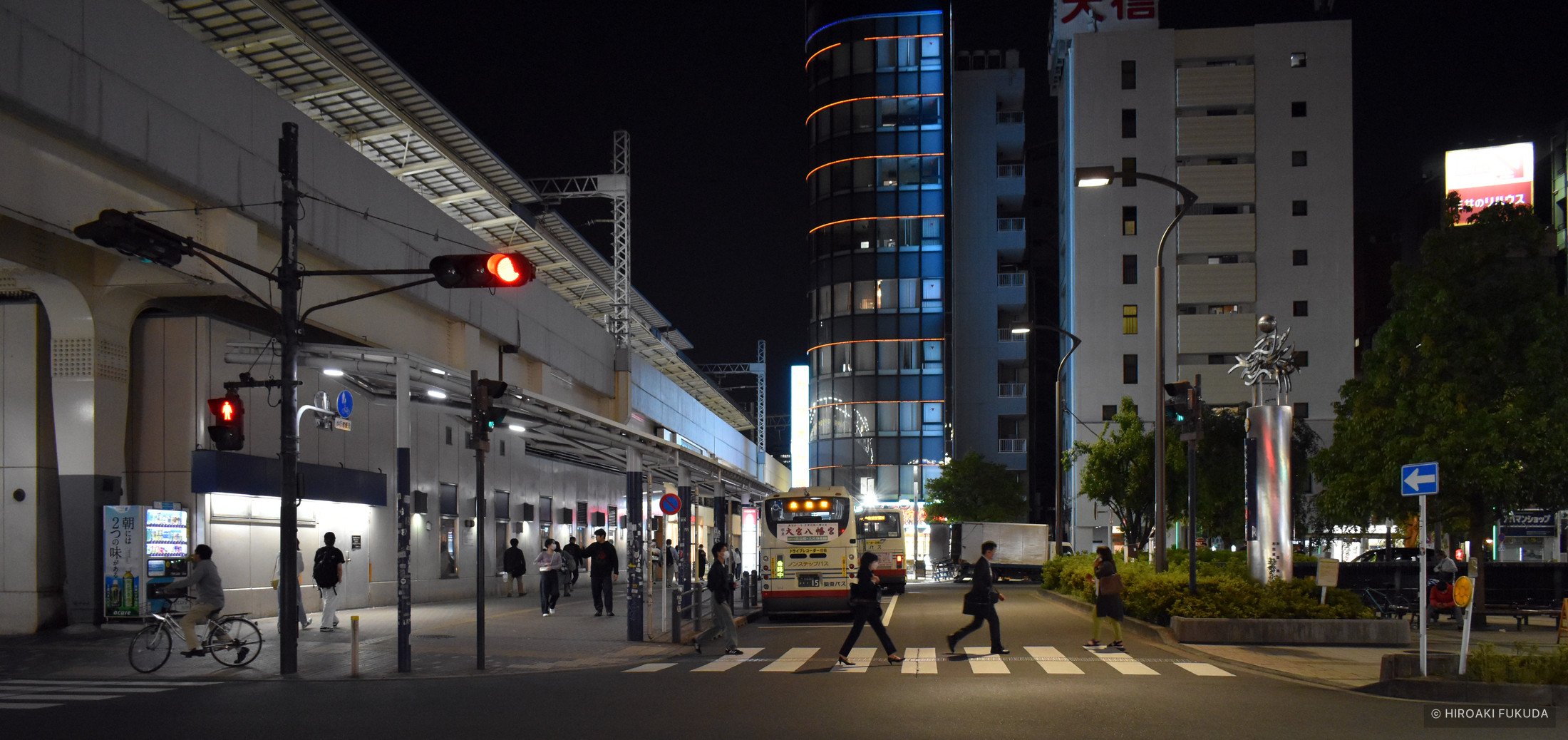 高架沿いの夜の交差点。赤い信号、バス、横断歩道を渡る人々が見える写真